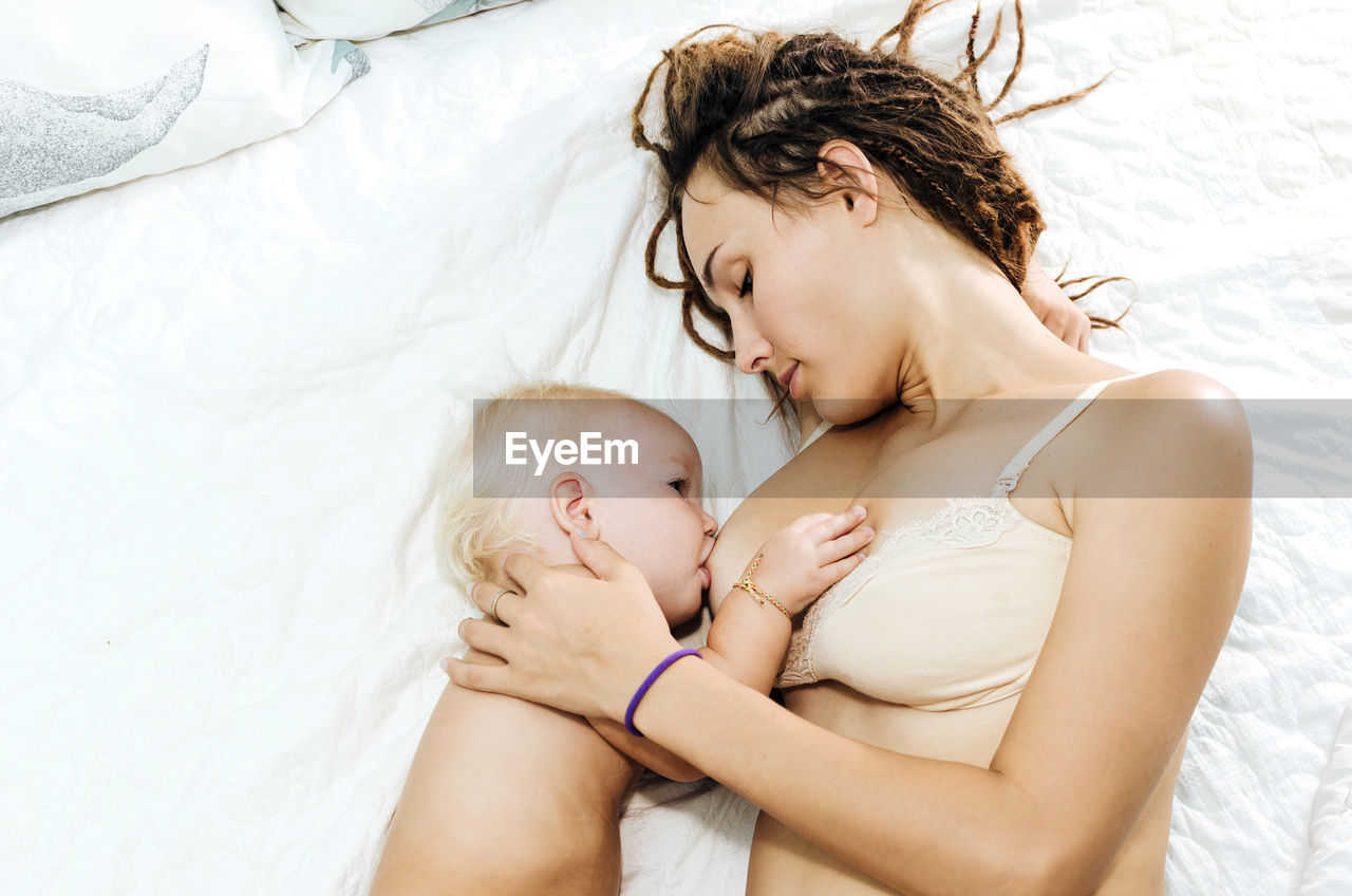 High angle view of mother breastfeeding shirtless daughter on bed at home