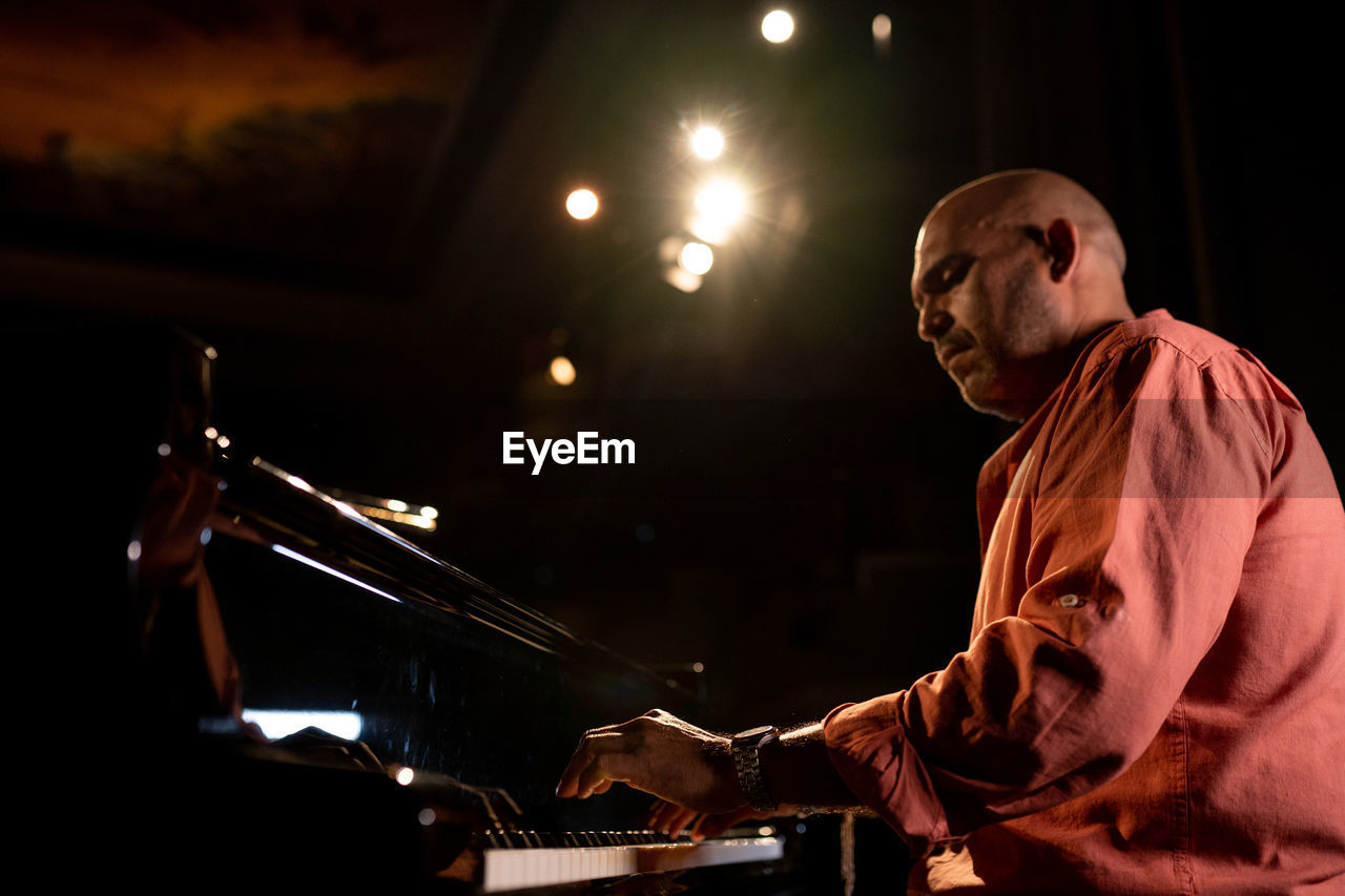 From below side view of bald male musician playing piano during performance on obscure stage in theater