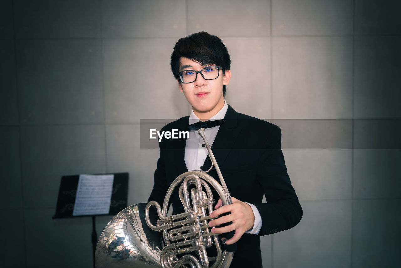 Portrait of man holding french horn while standing against wall