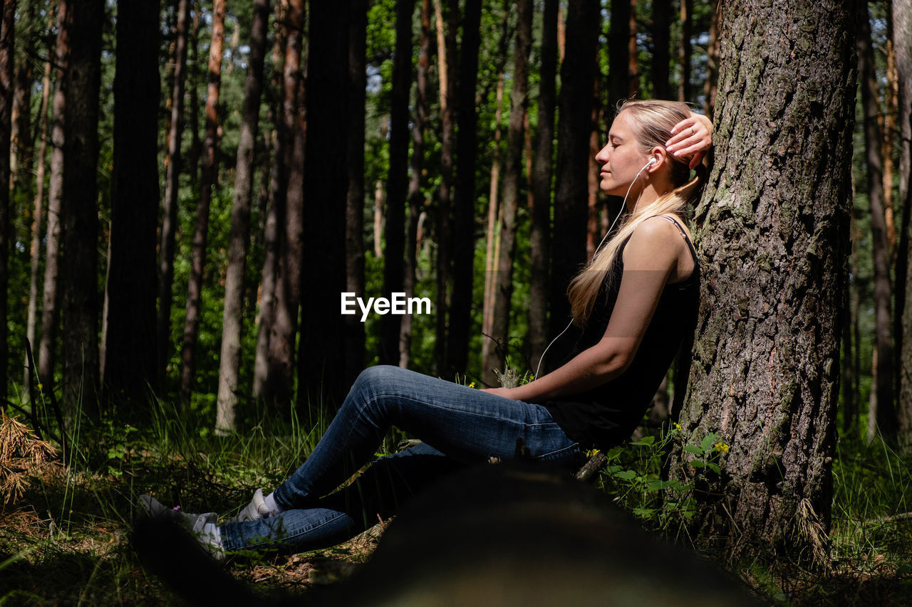 A girl in a forest sits leaning against a tree, with headphones listening to music. digital detox.