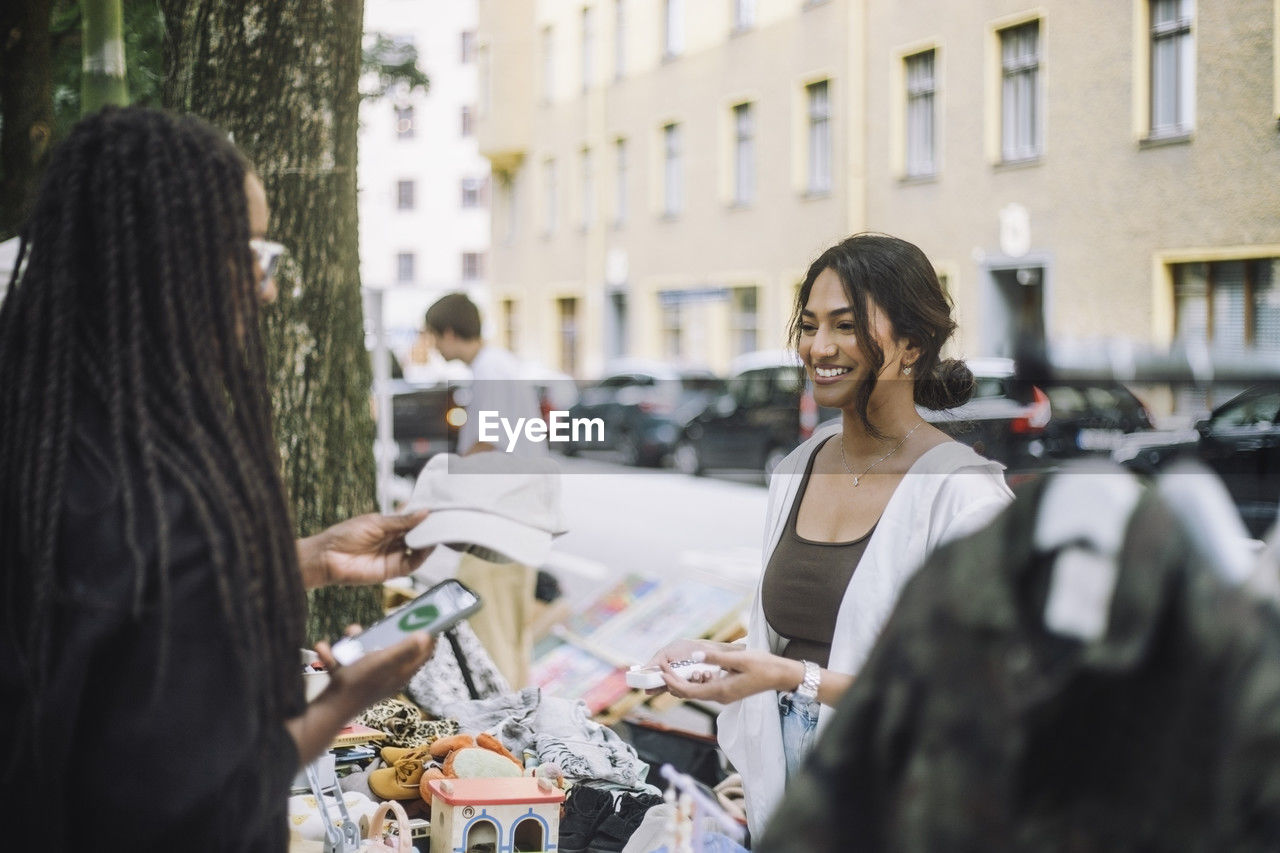 Woman buying cap from stall while talking to smiling female vendor at flea market