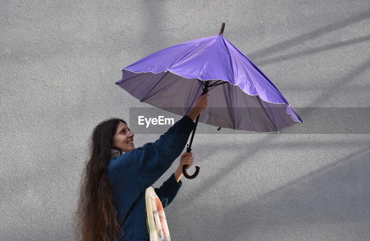 Side view of woman opening purple umbrella while standing against gray wall
