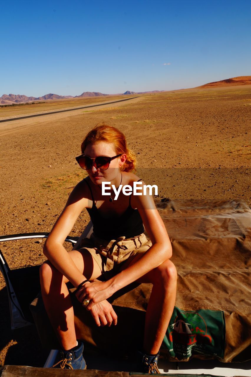 Young woman in sunglasses sitting at desert against clear sky