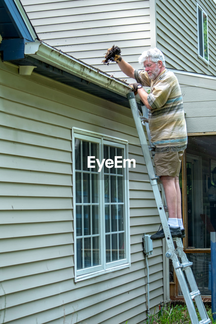 An older man carefully stands on a ladder and cleans leaves and debris from his home's gutters