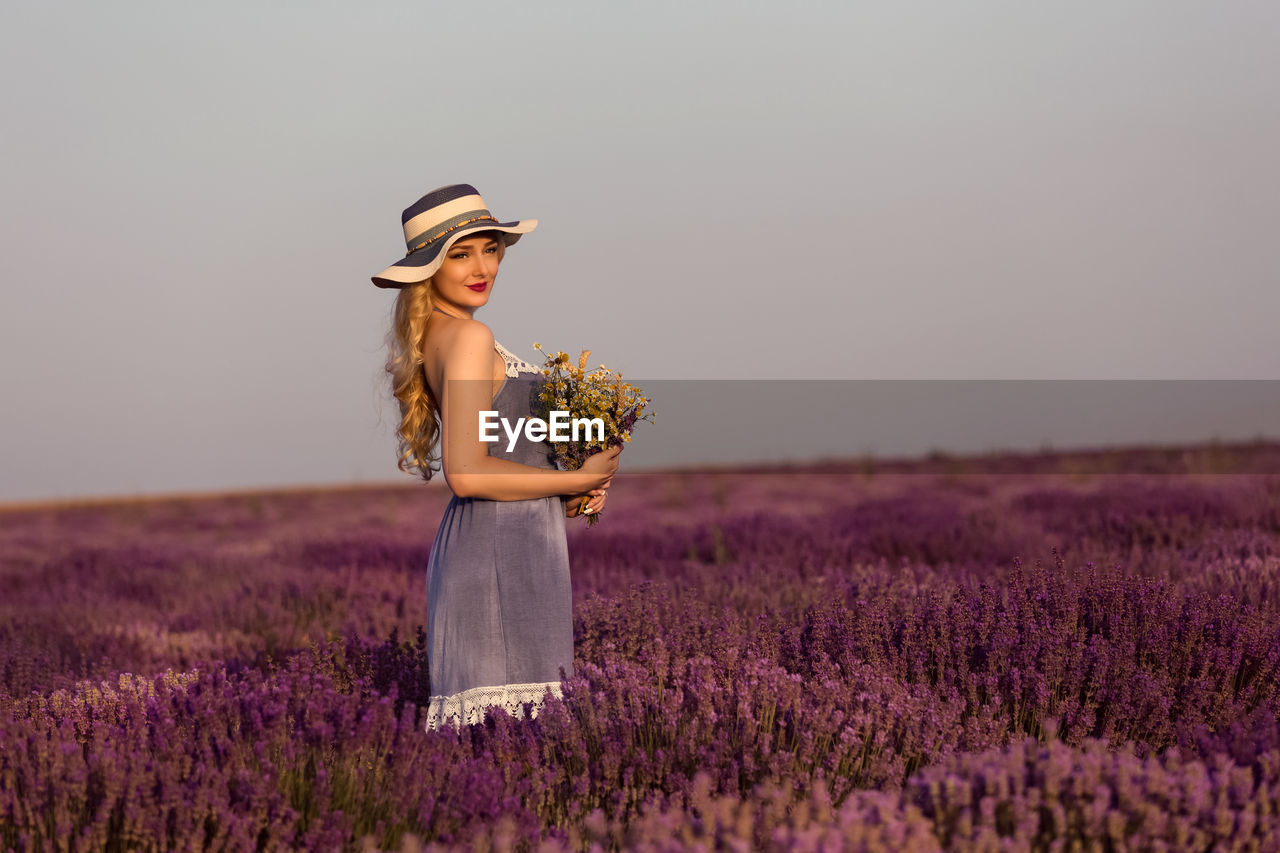Young woman holding bouquet of wild flowers in lavender field at sunset