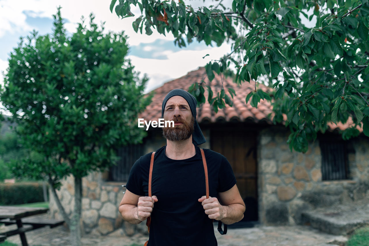 Calm male tourist with backpack standing near stone house in settlement and looking away