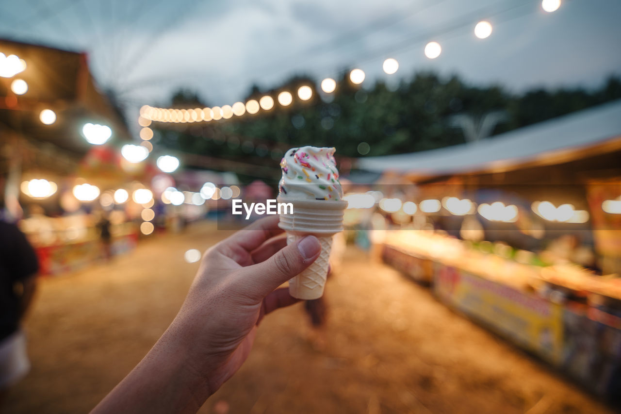 hand, holding, illuminated, night, focus on foreground, food and drink, one person, food, city, frozen, adult, architecture, sweet, ice cream cone, cone, sweet food, celebration, outdoors, personal perspective, lighting equipment, dairy, nature, leisure activity, dessert, cold temperature, frozen food, lifestyles, street