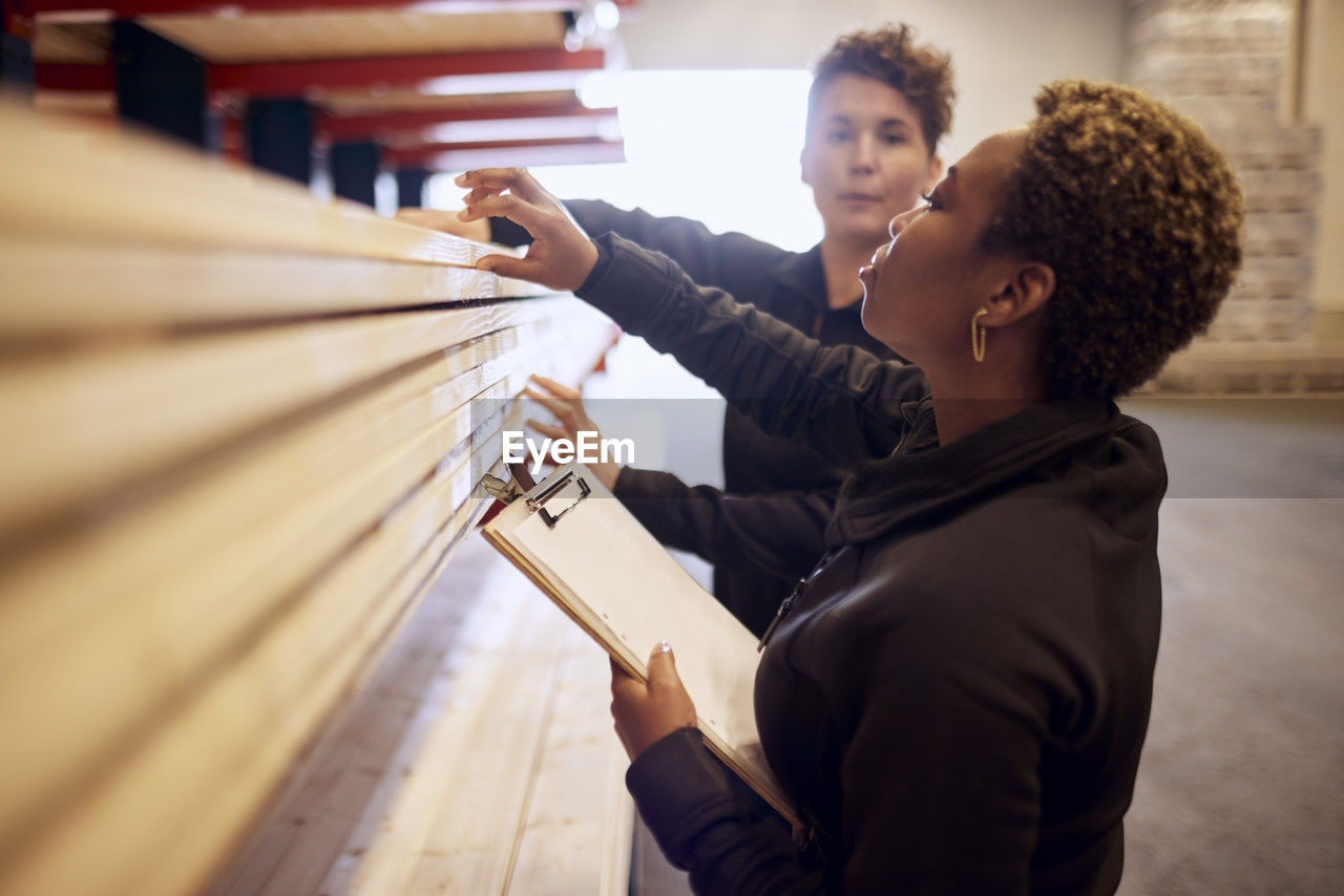 Multiracial female colleagues examining planks on rack in lumber industry