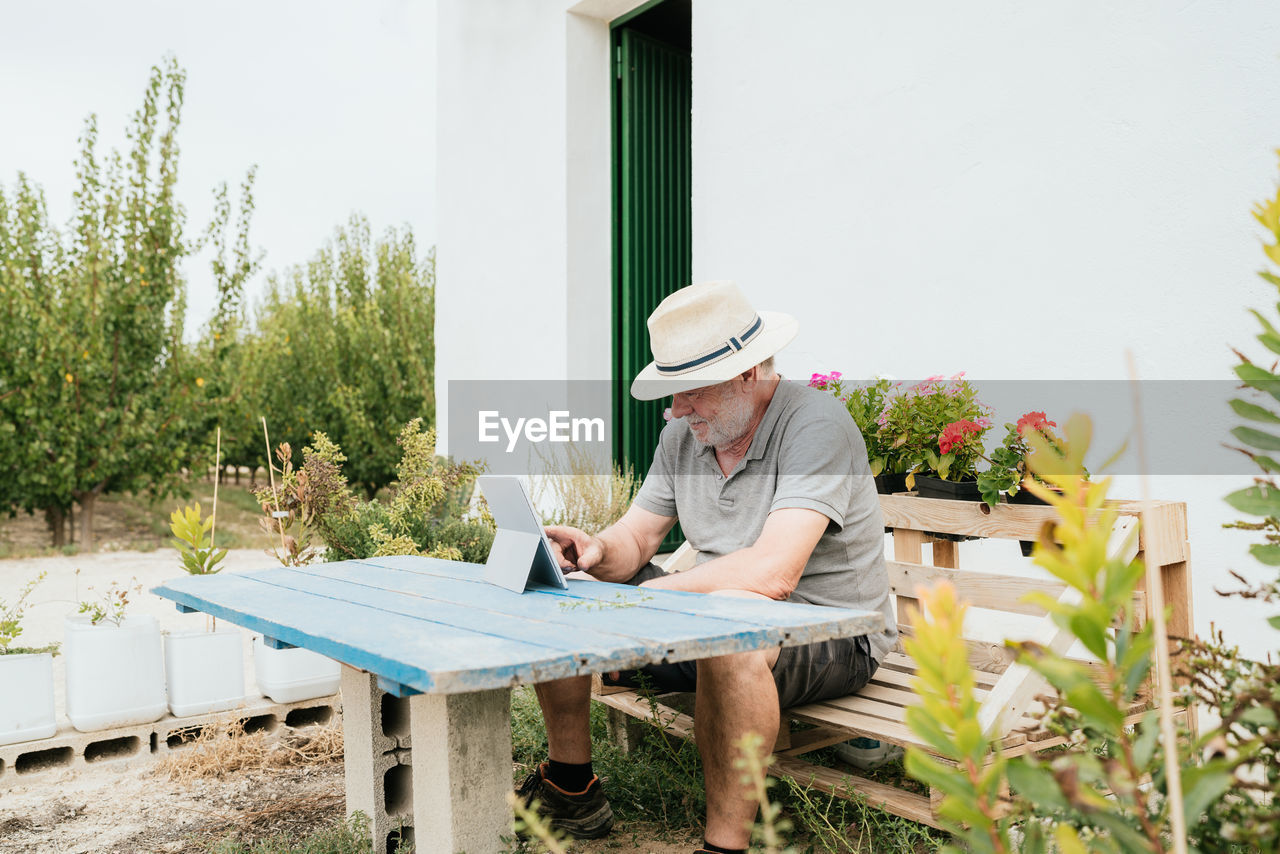 Elderly man in casual clothes and hat sitting on wooden bench at shabby table and browsing data on tablet during break in garden outside house on summer day in countryside