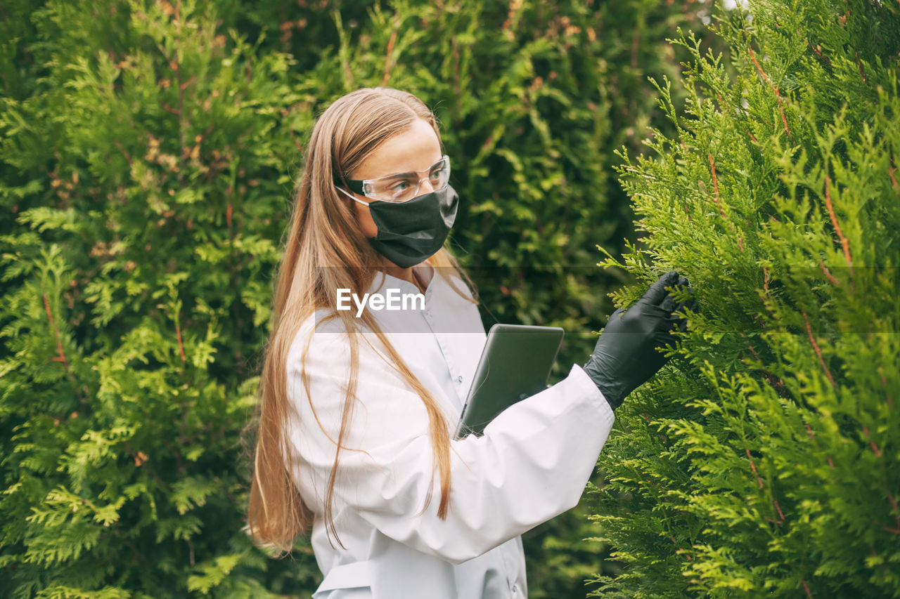 YOUNG WOMAN USING SMART PHONE WHILE STANDING ON PLANTS