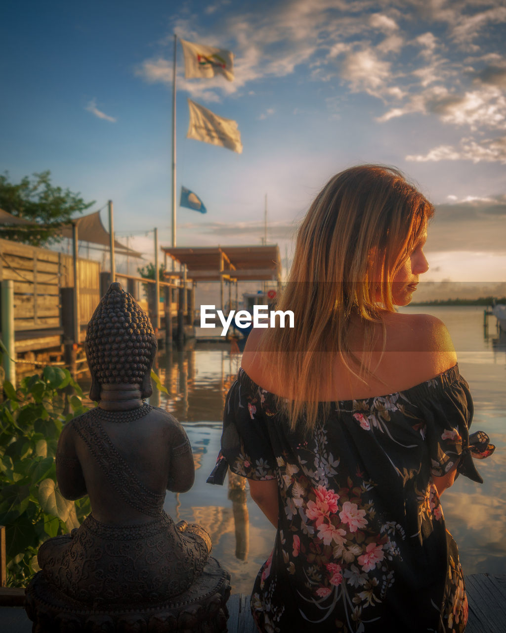 Rear view of woman sitting by buddha statue on shore against sky during sunset