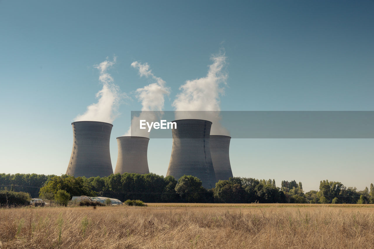 cooling tower, nuclear power plant, factory, power station, environment, power generation, sky, environmental issues, outdoor structure, smoke, pollution, industry, smoke stack, air pollution, nature, plant, building exterior, environmental damage, no people, built structure, day, emitting, landscape, fumes, clear sky, architecture, electricity, outdoors, tree, fossil fuel, field, cloud, climate change, wind, social issues, land