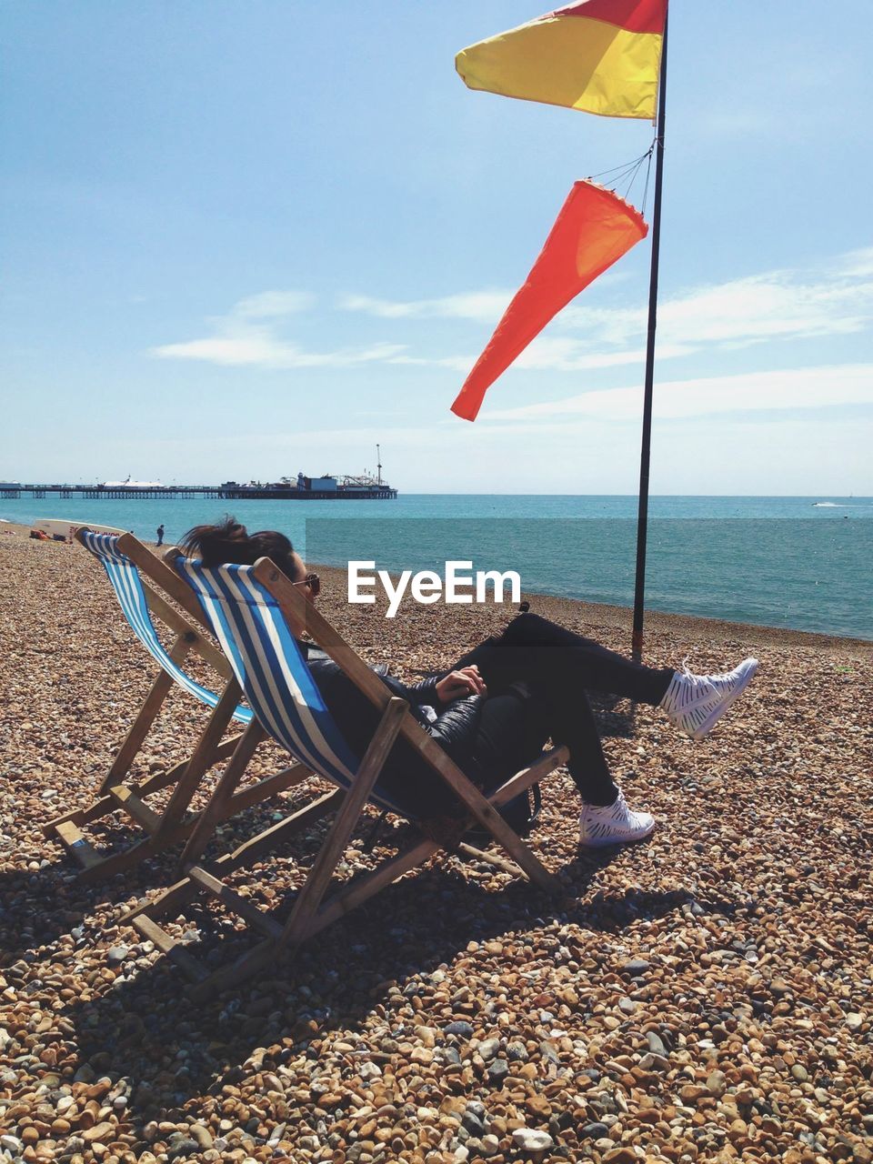 Side view of mature woman sitting on deck chair at beach against sky during sunny day