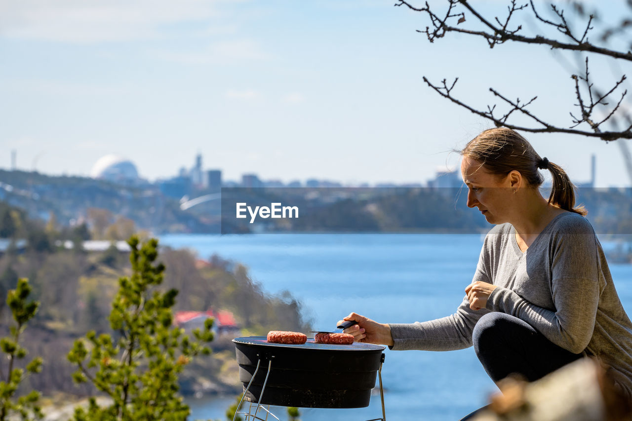 Side view of woman preparing meat on barbecue by river