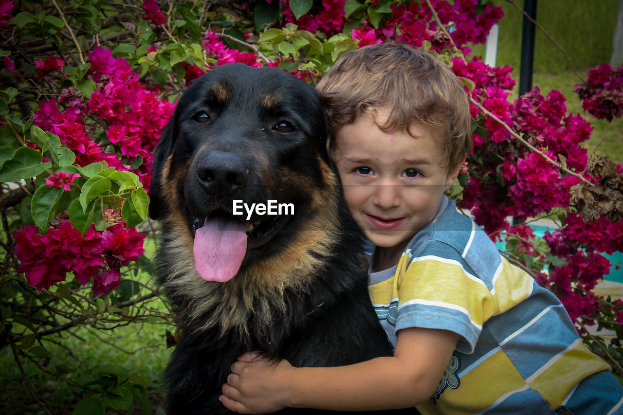 Portrait of smiling boy embracing dog against bougainvillea in yard