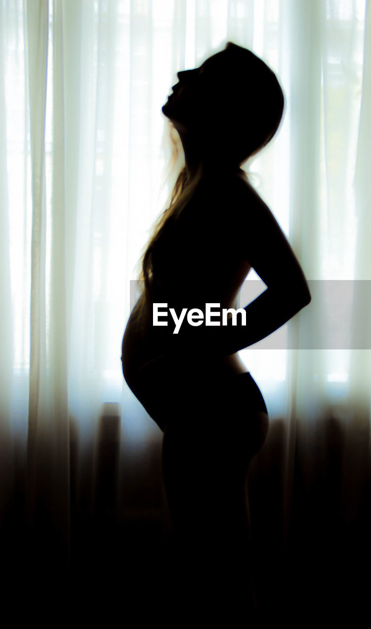 Side view of sensuous pregnant woman standing by curtain at home