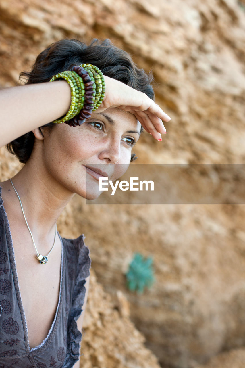 Close-up of smiling woman shielding eyes while looking away against rock