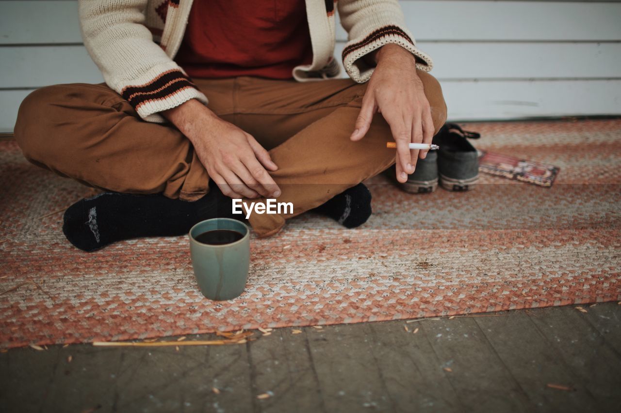 Low section of man holding cigarette while sitting by coffee mug at sidewalk