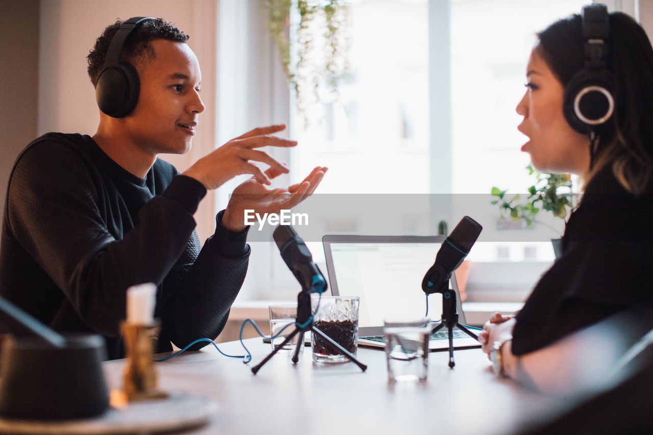 Male blogger gesturing to female wearing headphones sitting at table