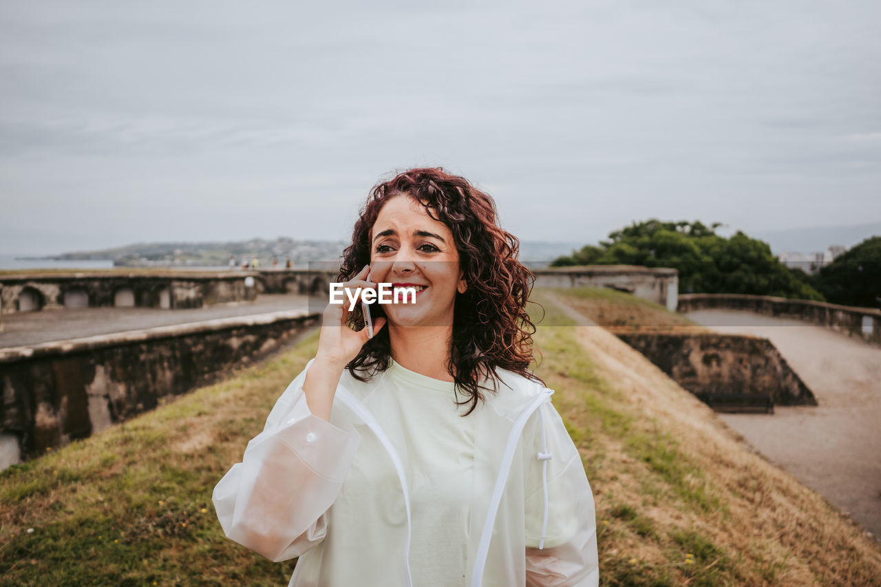 Smiling woman talking over phone while standing on grass against sky