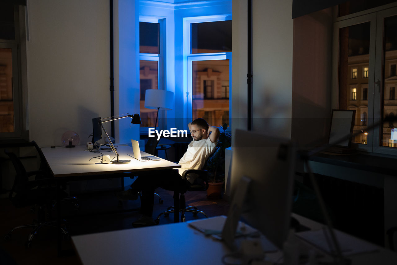 Happy calm man look at laptop hold hands behind head in night office satisfied with finished work