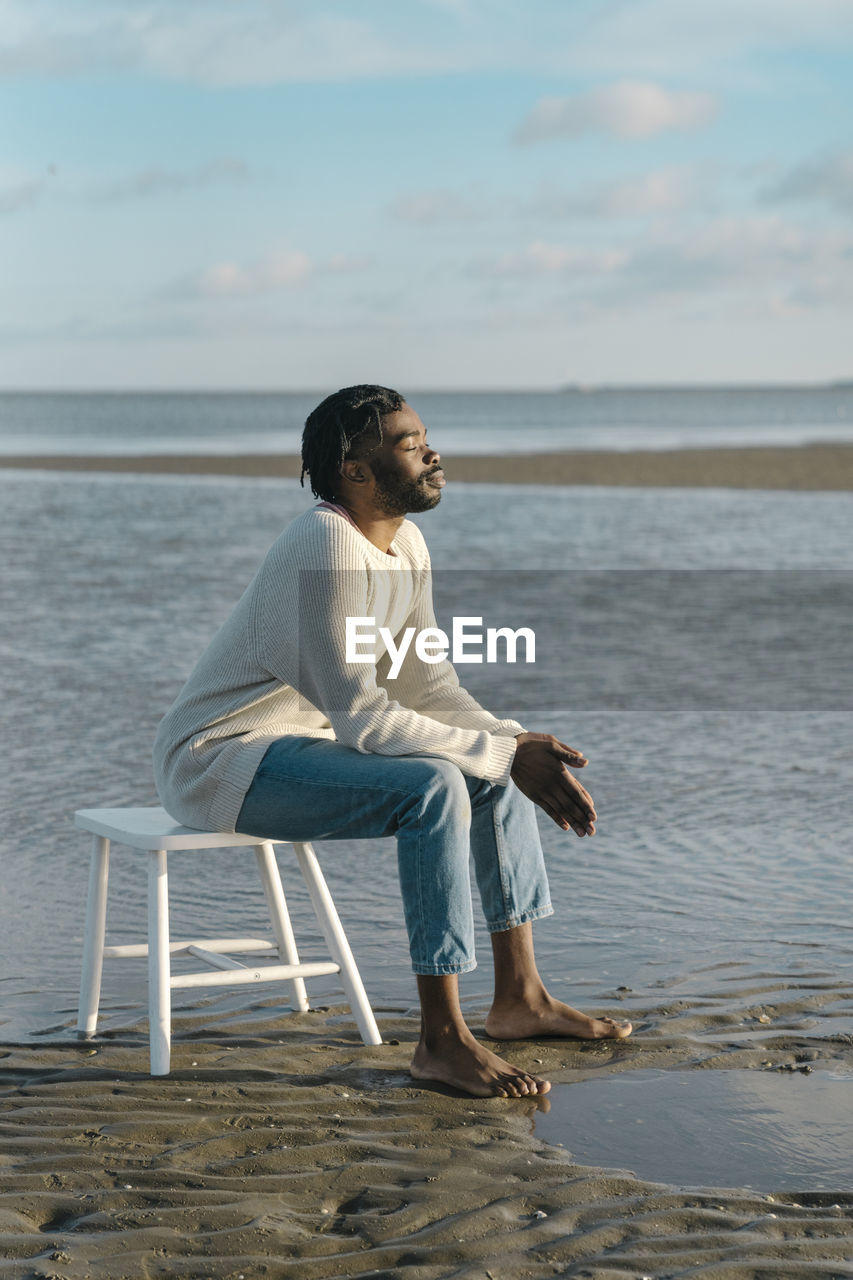 Relaxed young man with eyes closed sitting on white stool at beach against sky