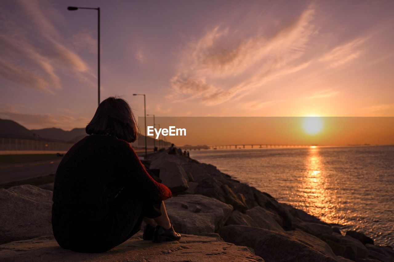 Side view of woman sitting at beach against sky during sunset