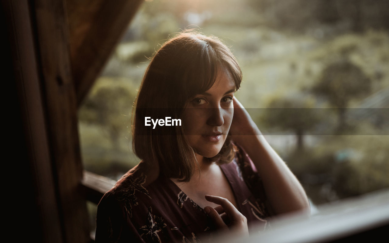 Portrait of young woman on ledge of wooden cabin at sunset