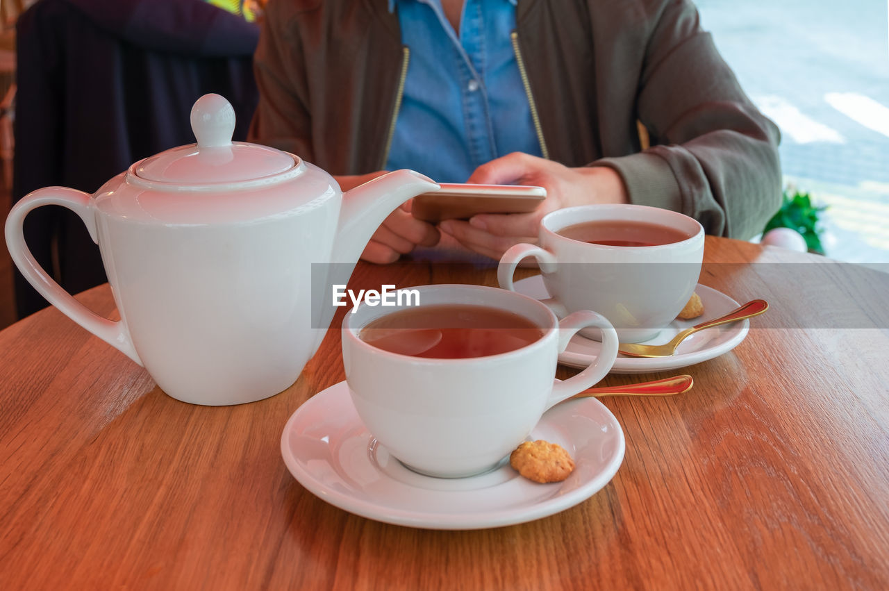 A teapot and two cups of tea on a table in a cafe. young woman holding a smartphone. 