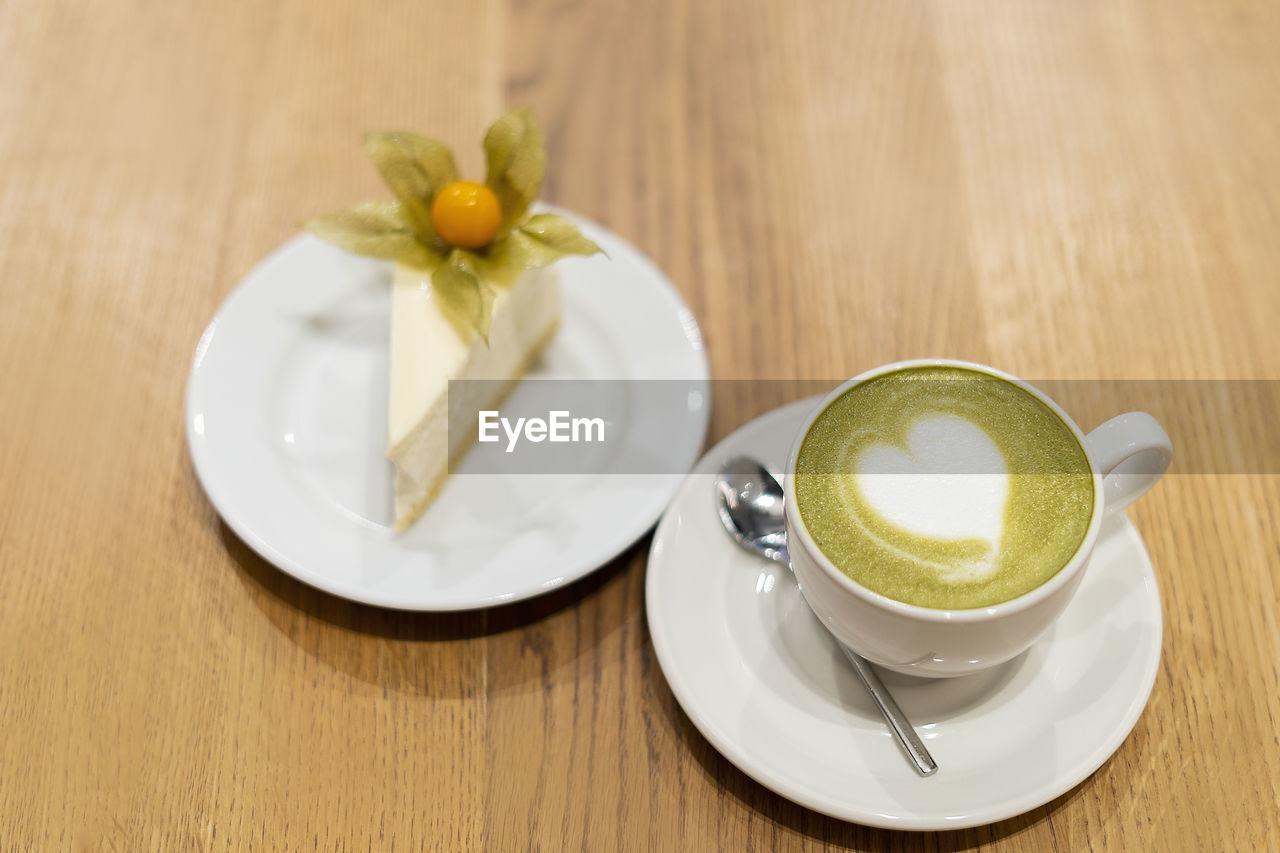 On the wooden surface stands a white cup of matcha tea. on a saucer is a white cheesecake with 