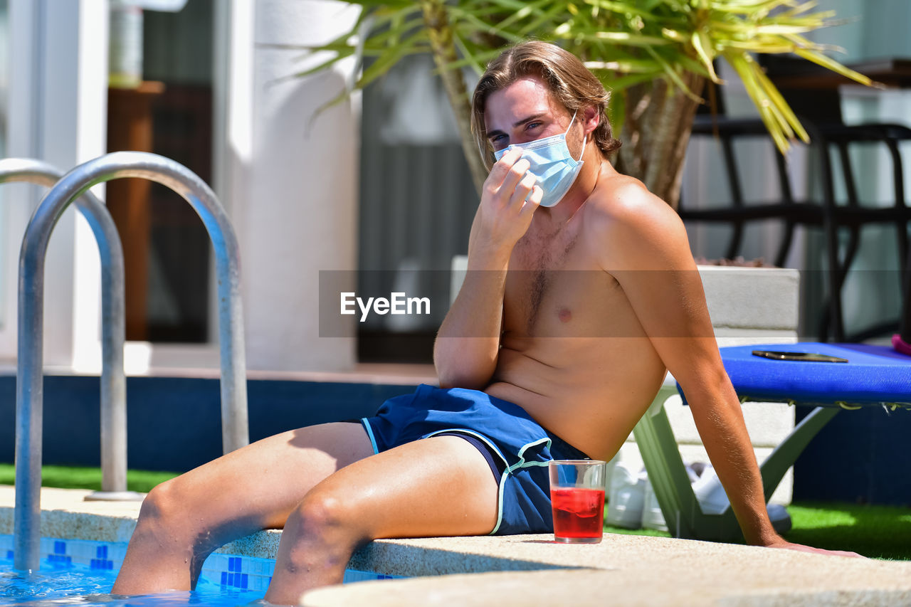 Shirtless man with protective face mask sitting in poolside of swimming pool