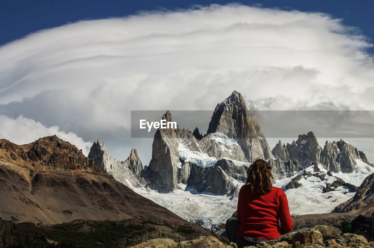 Rear view of woman sitting on rock against cloudy sky