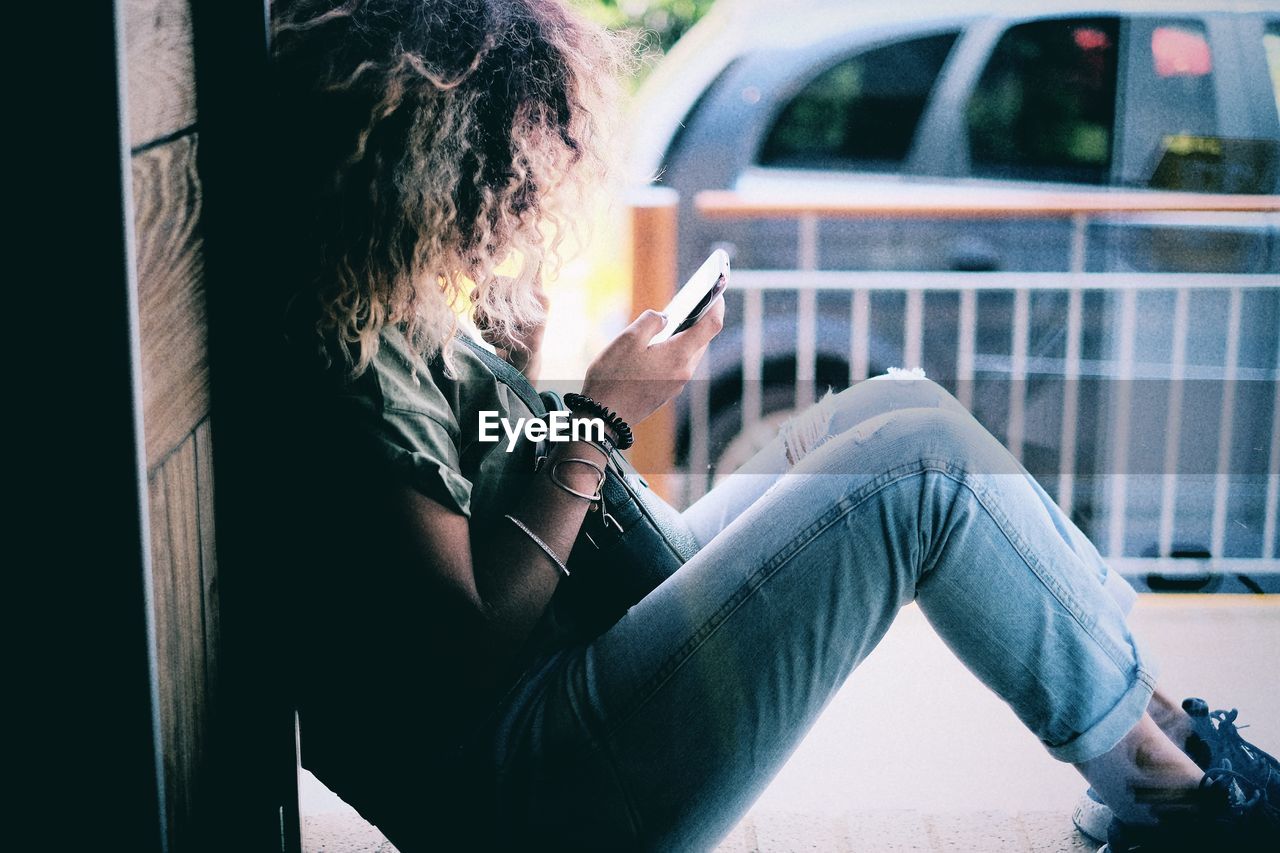 Side view of woman using cellphone while sitting on window