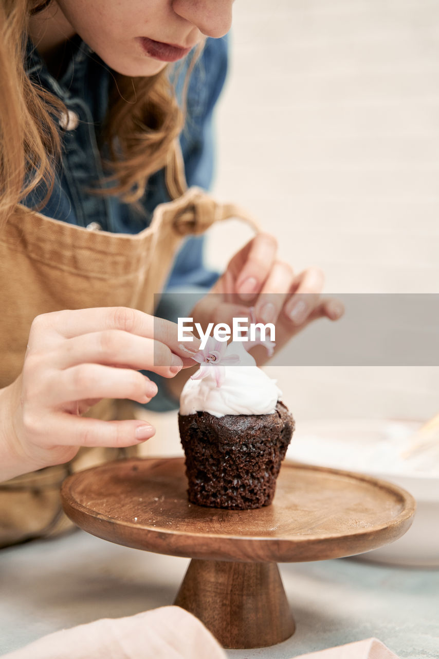 Crop young female decorating chocolate cupcake with sweet whipped cream and flowers on cake stand while cooking at home