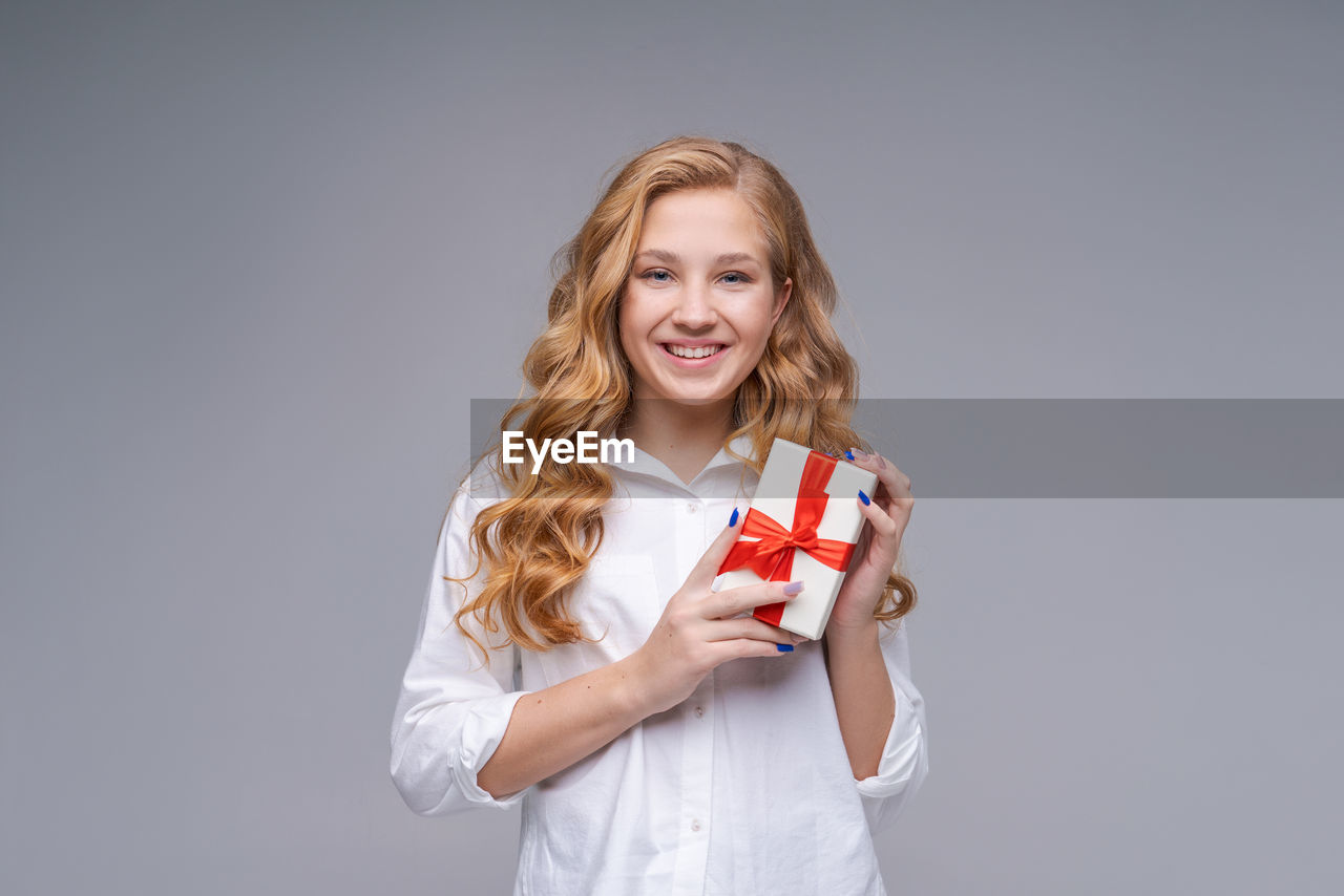 studio shot, smiling, happiness, one person, portrait, emotion, women, indoors, looking at camera, blond hair, gray background, gray, holding, cheerful, long hair, positive emotion, child, waist up, front view, copy space, person, standing, hairstyle, childhood, smile, teeth, finger, adult, female, casual clothing, photo shoot, clothing, colored background, cut out, hand, young adult, cute, human face, fun, white background