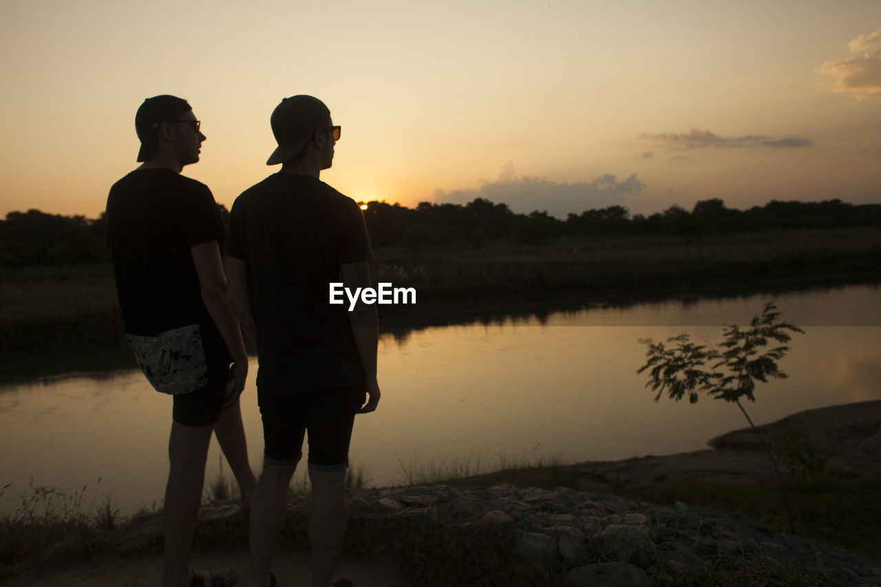 REAR VIEW OF SILHOUETTE FRIENDS STANDING BY LAKE AGAINST SKY