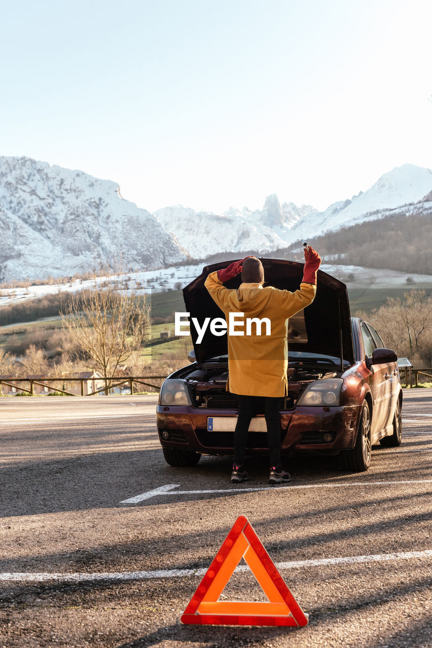 Back view full length person in outerwear opening car hood parked near warning triangle road sign in sunny winter highlands person