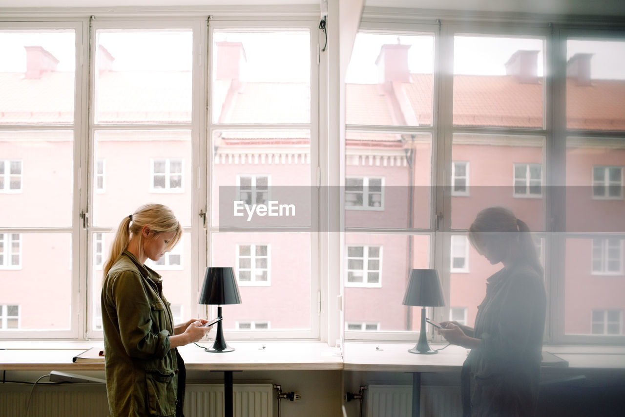 Businesswoman using smart phone while reflecting on glass at office