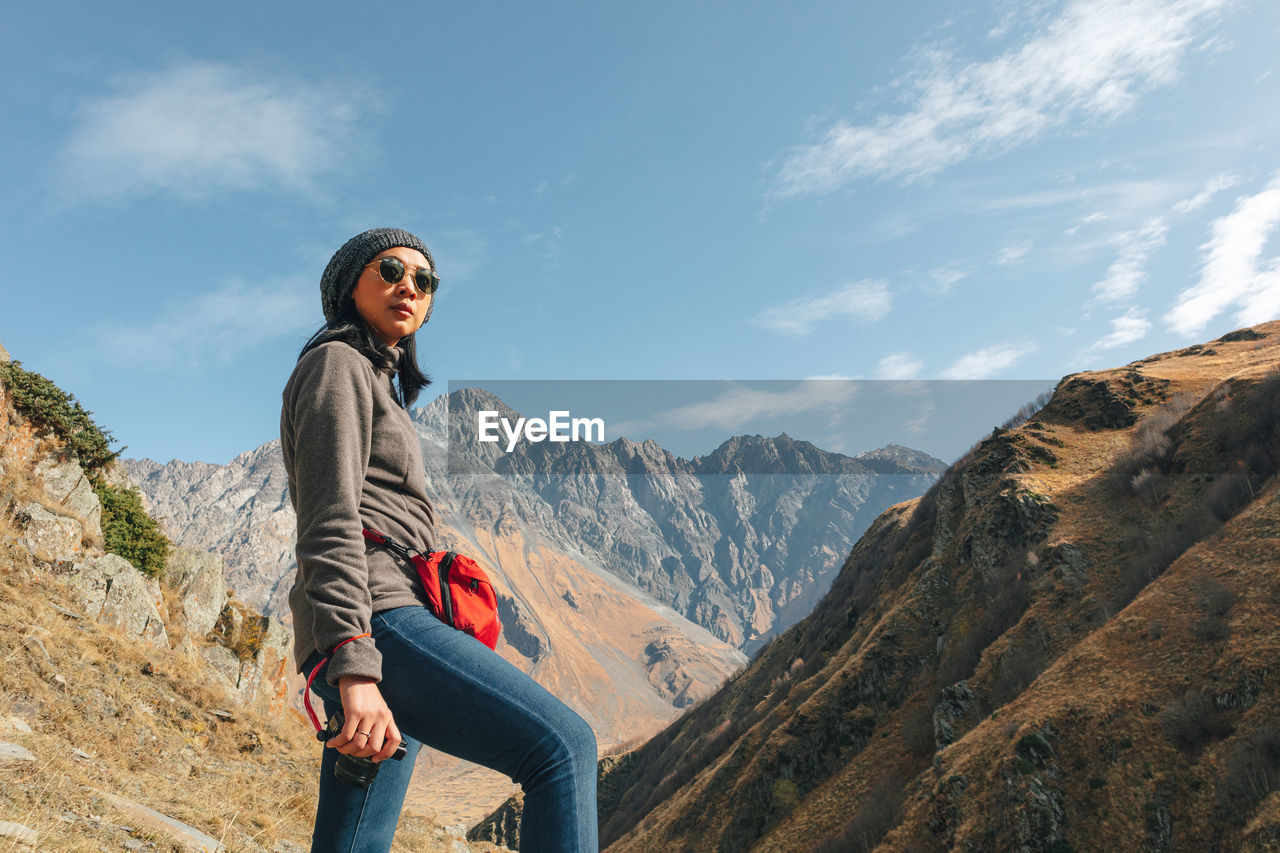 Low angle view of woman wearing sunglasses standing on mountain against sky