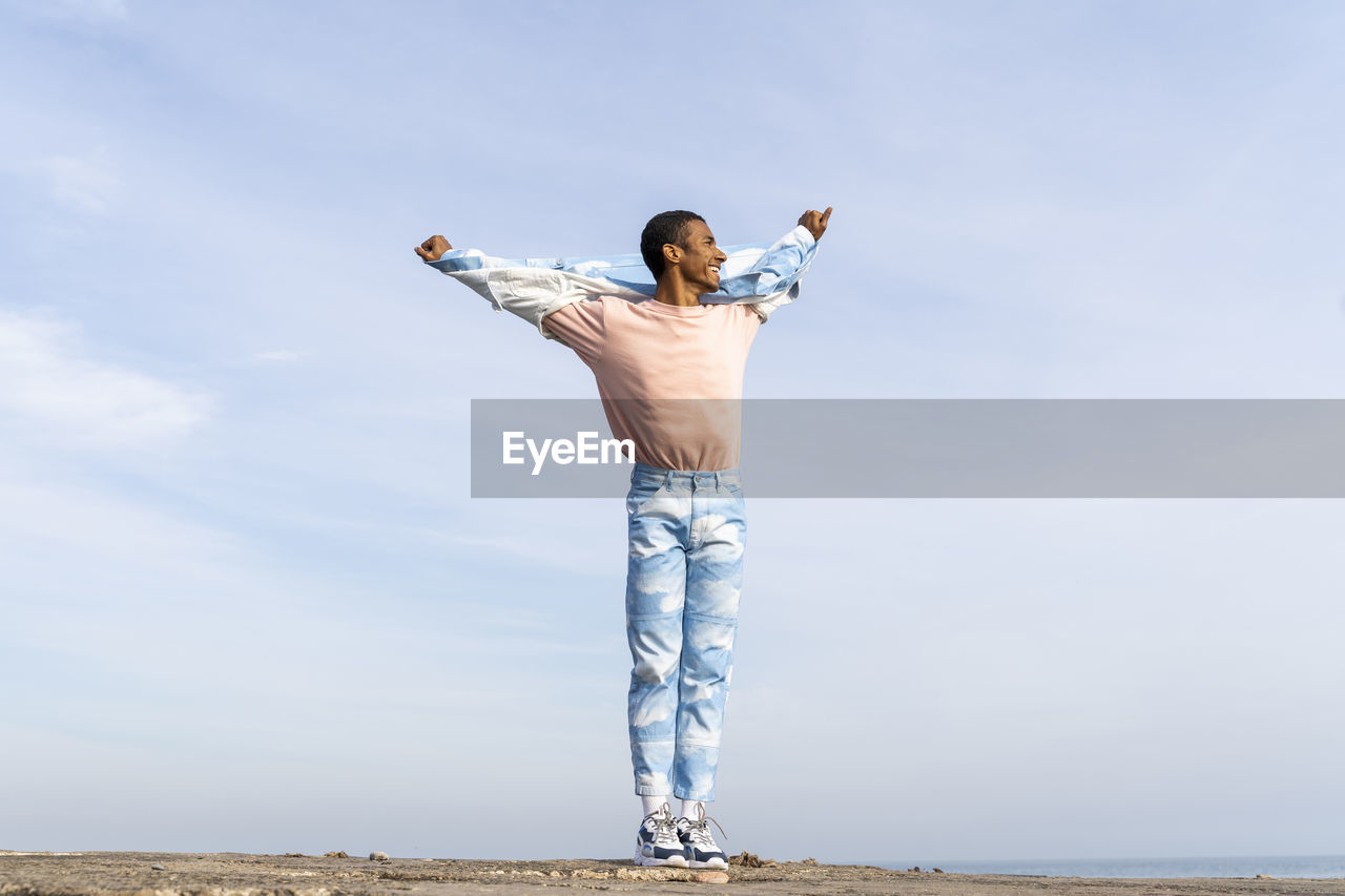 Smiling man standing with arms outstretched against sky
