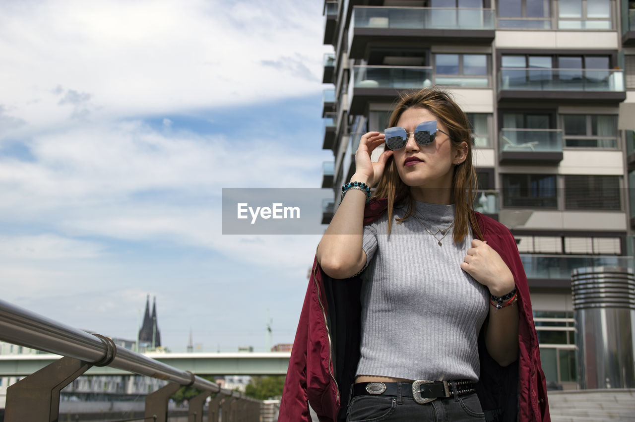 Confident young woman standing against cloudy sky in city