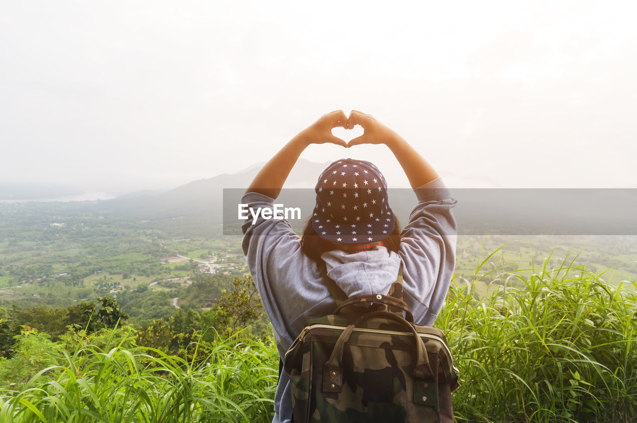 Rear view of woman gesturing heart shape while standing on mountain against sky during foggy weather