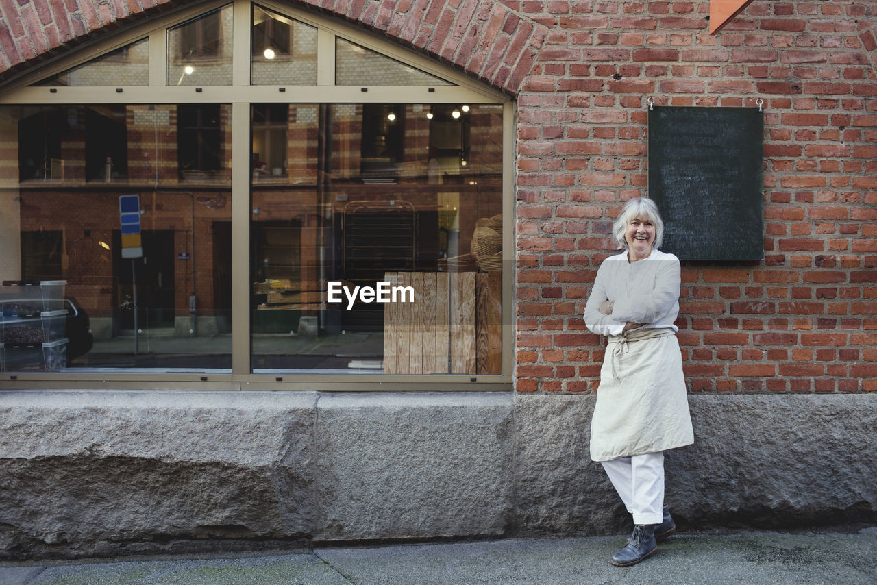 Full length of smiling baker standing on sidewalk against brick wall