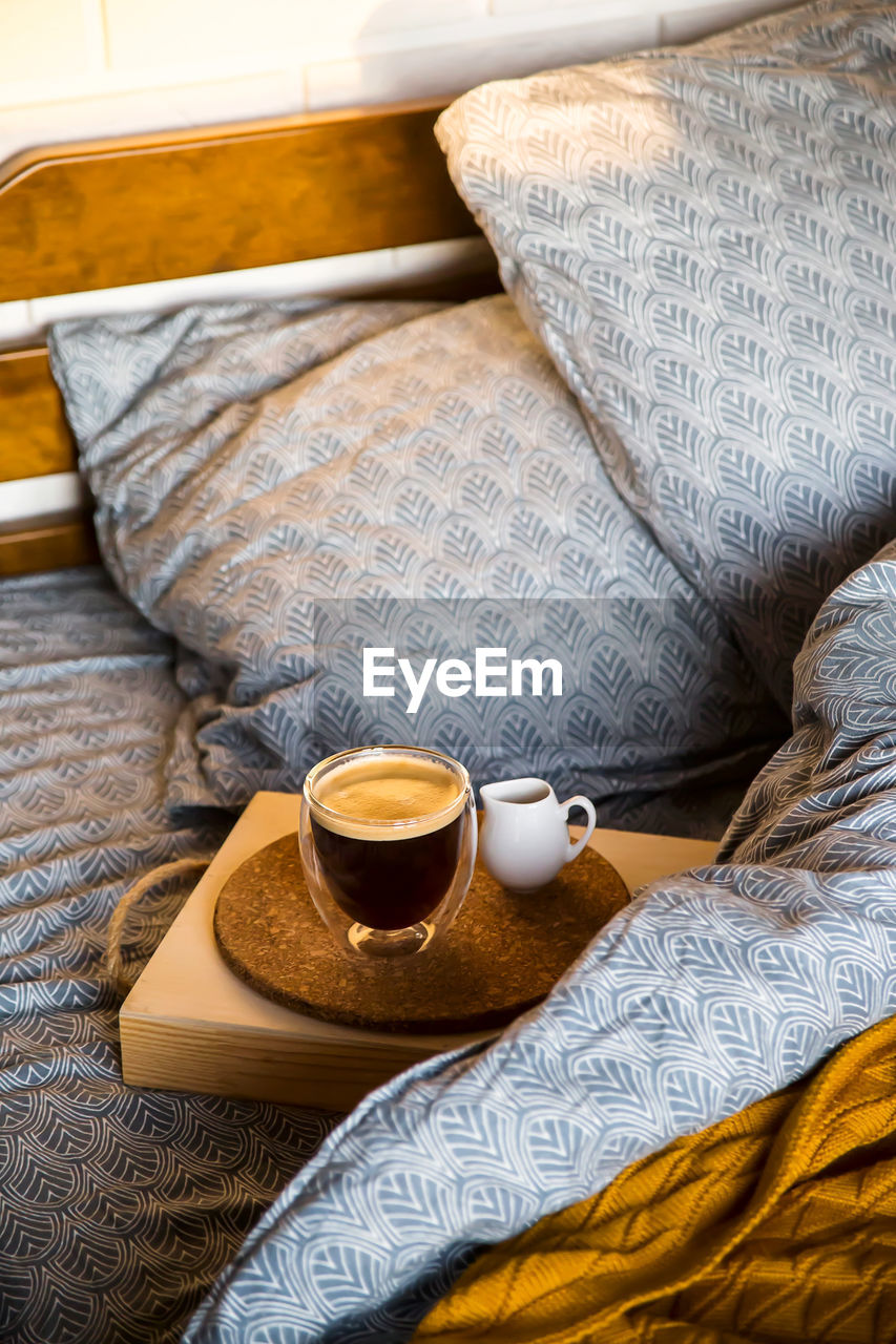 Coffee in a modern cup with a double bottom on a tray with creamer in bed with blue textured linen.