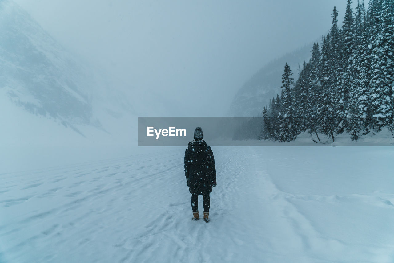 Female in winter clothes stands in snow field looking at pine forest and snow-capped mountains.