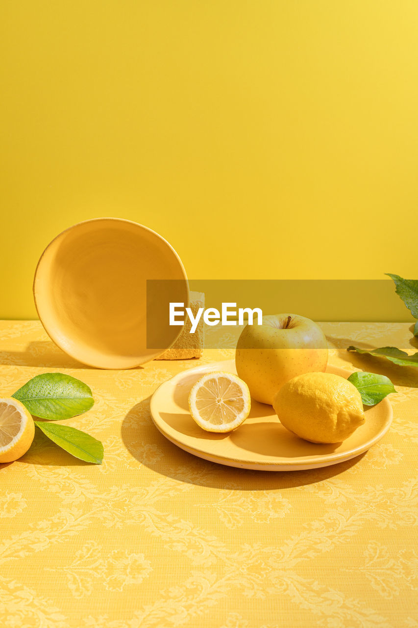 Fresh whole and halved sour lemons near apple on vivid ceramic plate on table on yellow background