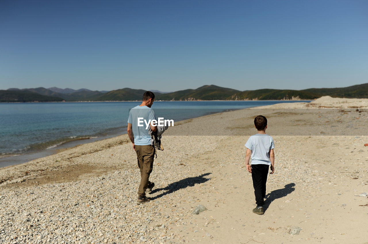 Man and preschool boy in blue identical t-shirts walk side by side on huge deserted beach and sea