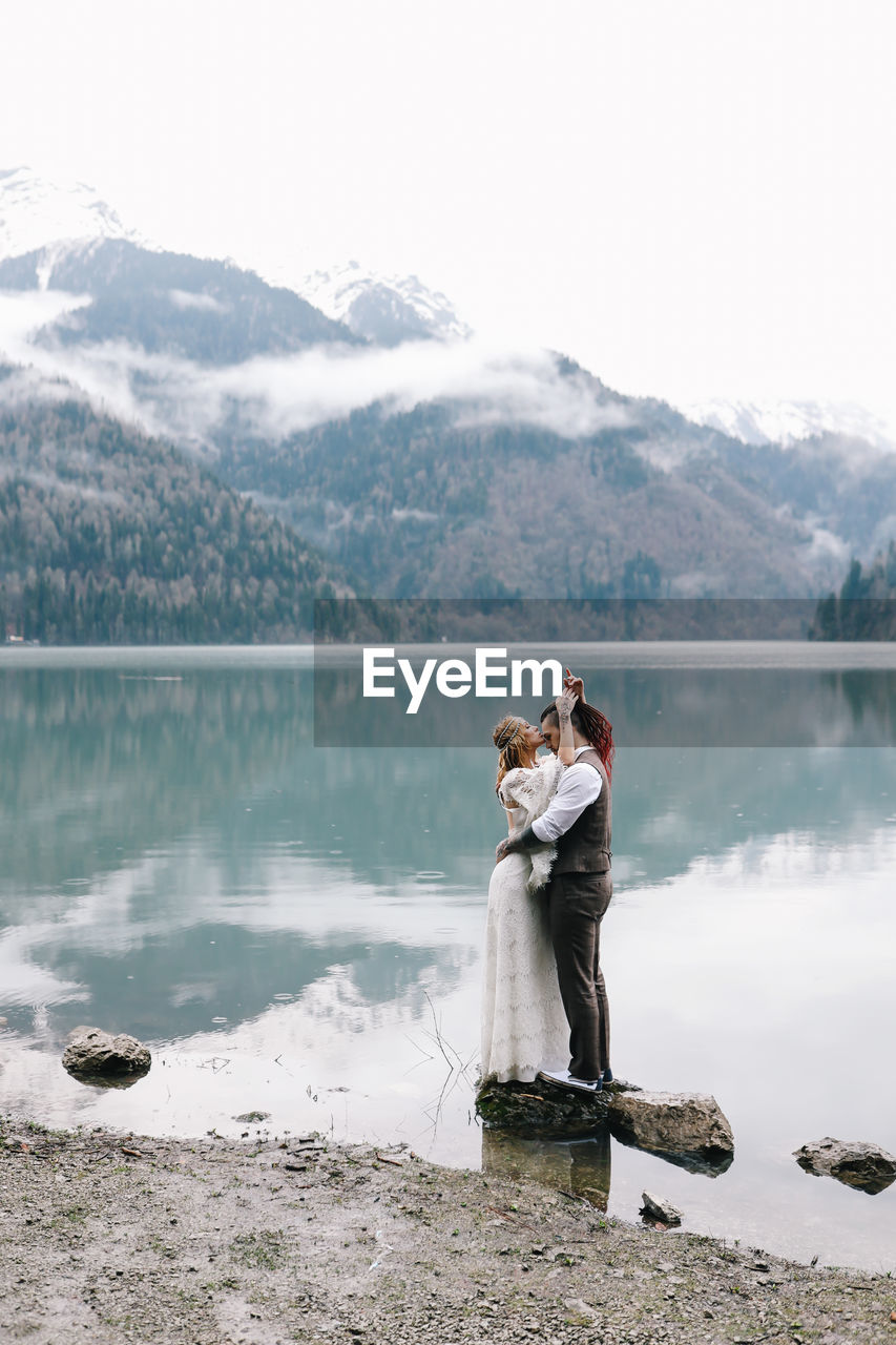 WOMAN STANDING BY LAKE AGAINST MOUNTAIN