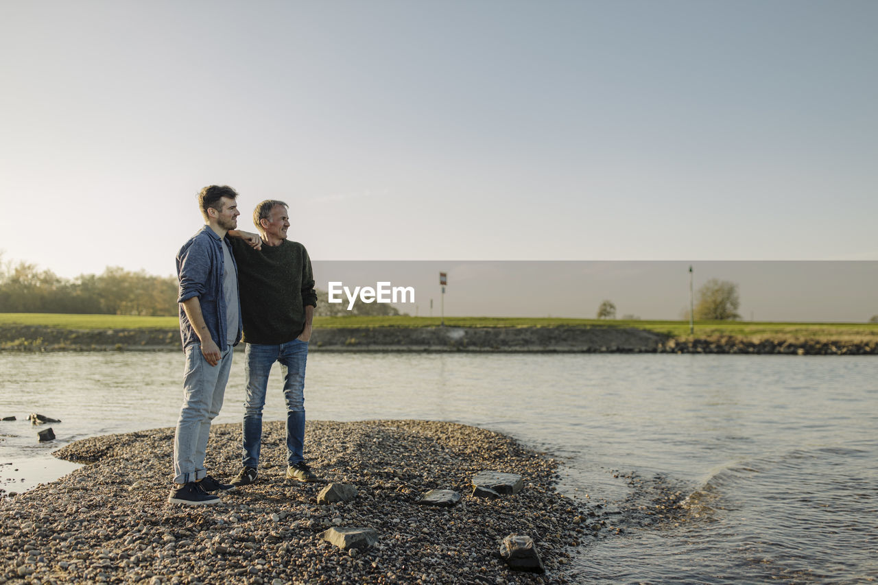 Son and father looking away while standing against clear sky at riverbank