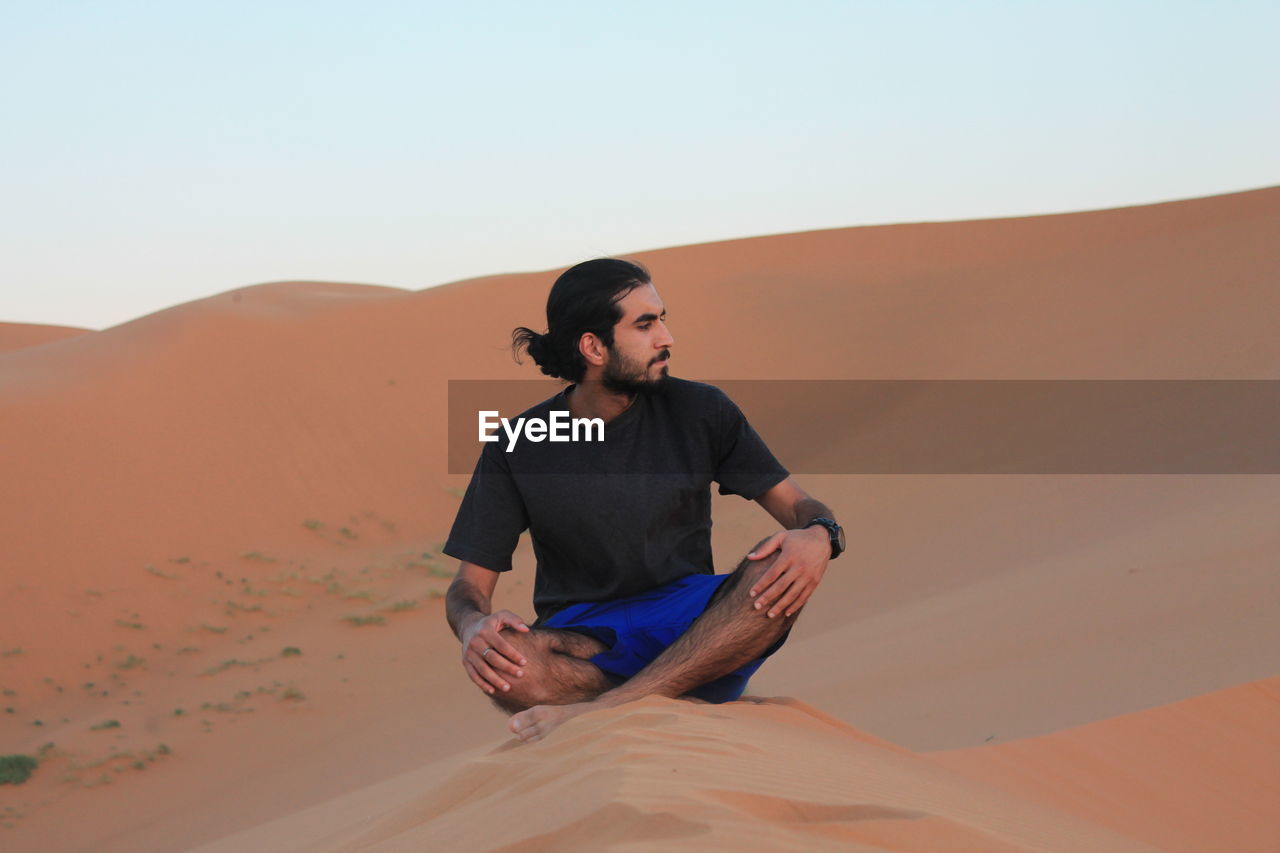 Man sitting on sand dune against clear sky